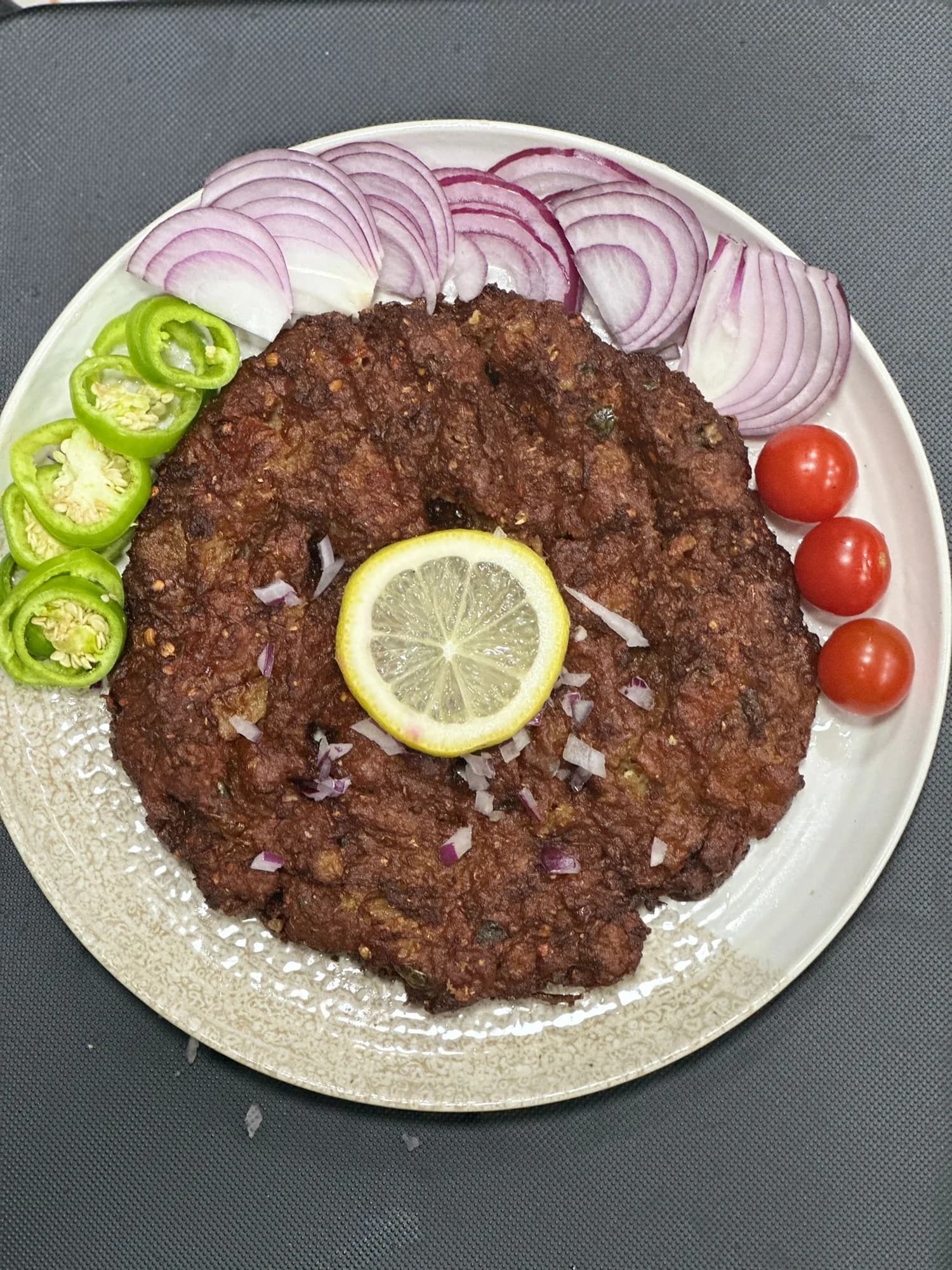 A chapli kebab served with green chillies, sliced red onion, cherry tomatoes and lemon